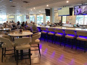 A bar area with round tables and chairs, a counter with a TV screen, and a view of the outside through large windows. - Lovely 2 bedroom and den house sleeping 6. Quiet with many great amenities. (North Fort Myers)