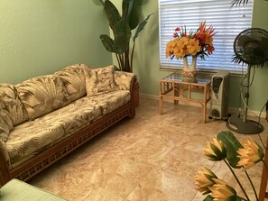 Un salon avec un canapé, une petite table sur laquelle est posé un vase de fleurs, un ventilateur et une grande plante.