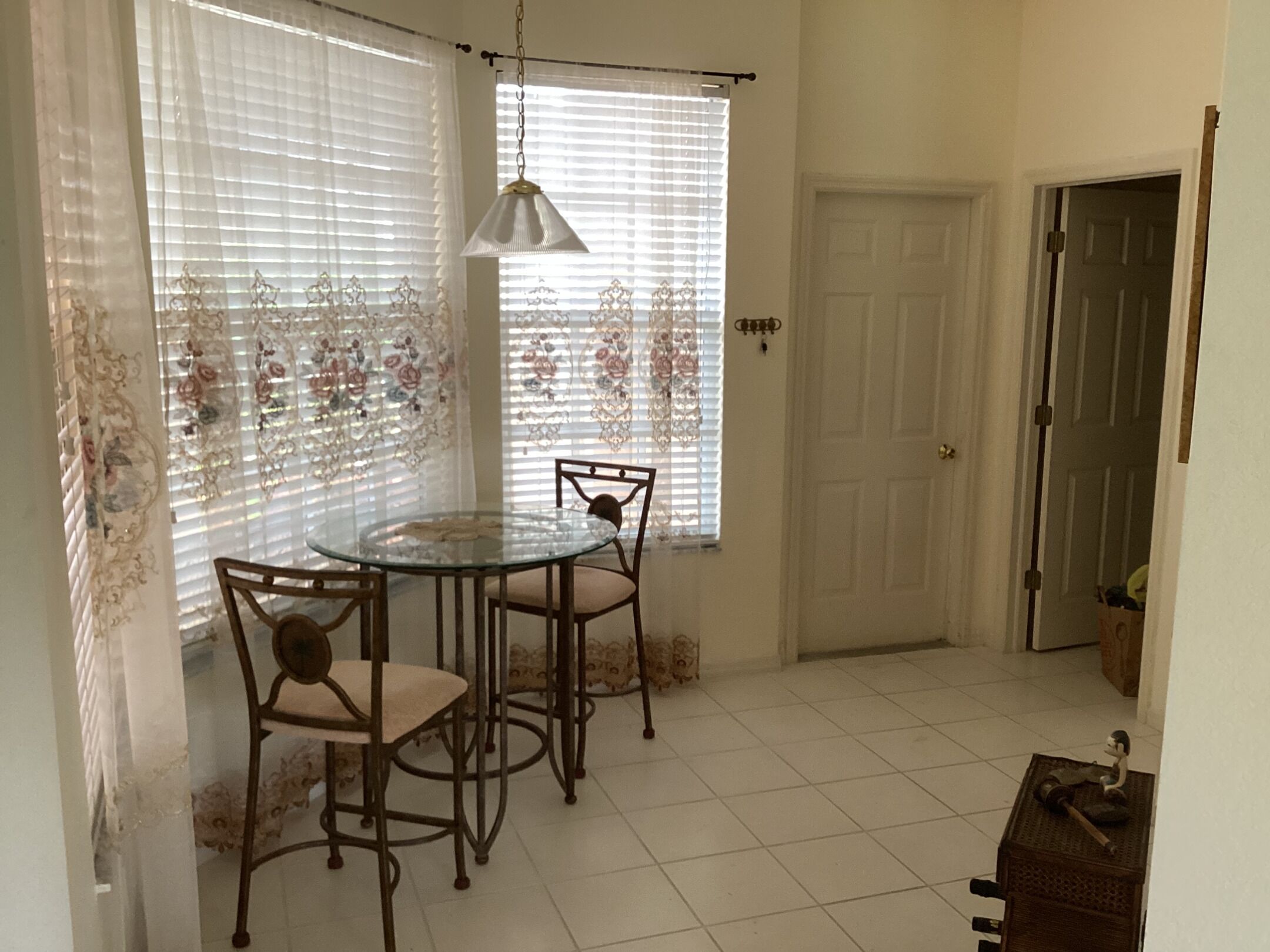 A dining area with a round glass table and two chairs, a door, and a window with decorative curtains.