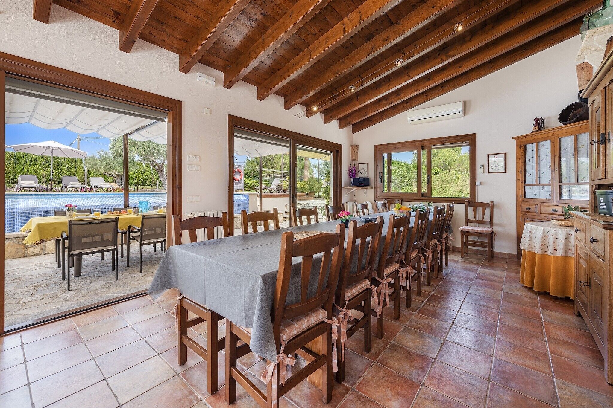 A dining area with a long table and chairs, a kitchen area with wooden cabinets, and a view of a swimming pool outside.
