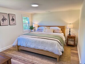 A bedroom with a large bed, two bedside tables with lamps, a window with a view of greenery, and two framed pictures on the wall. - The Little House on Rocky Hill - Peaceful 2BR Newberg Wine Country Retreat (Newberg)