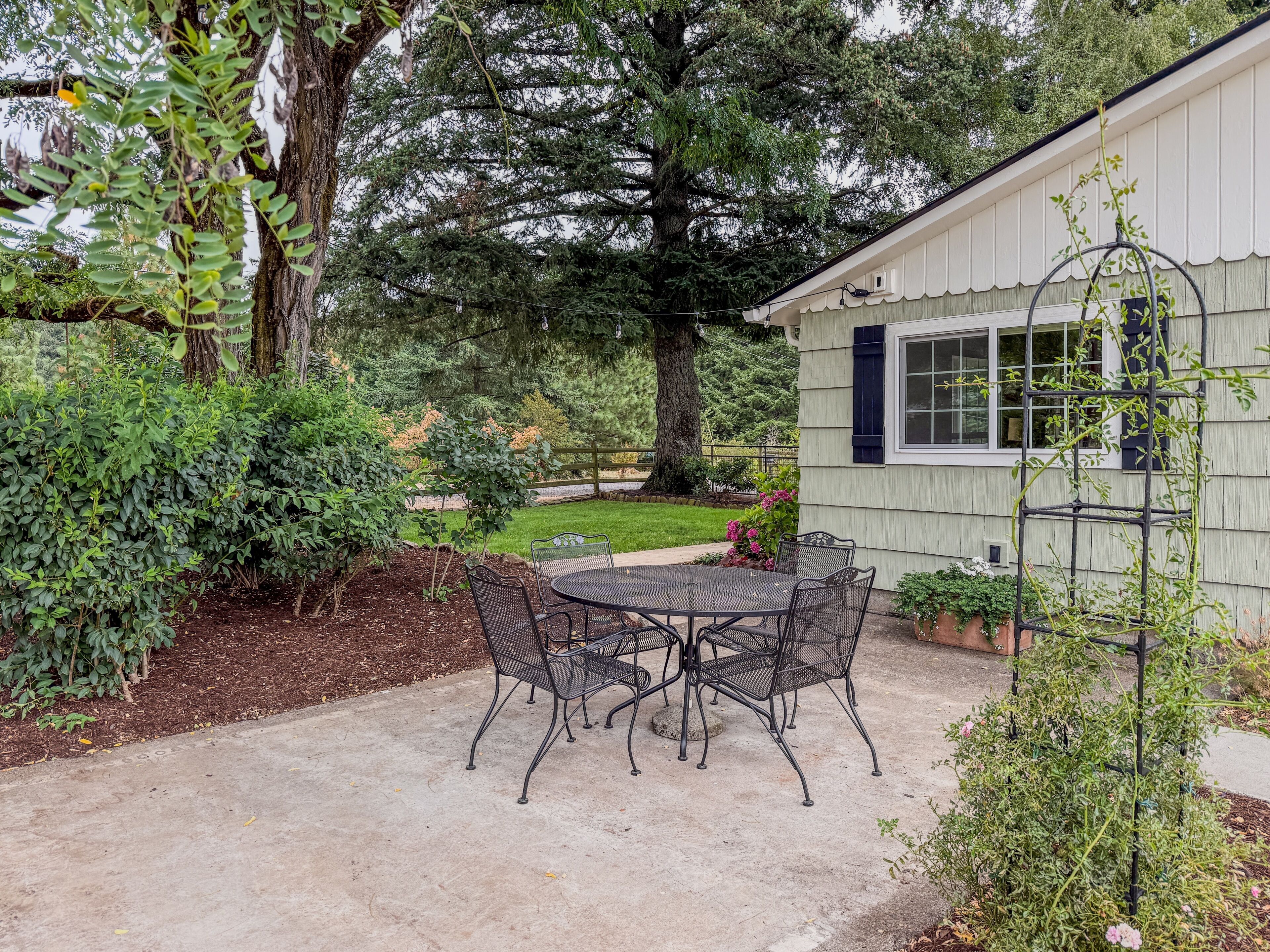 A patio with a table and chairs, surrounded by greenery and a building with a window.