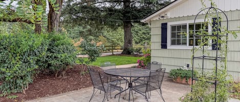 A patio with a table and chairs, surrounded by greenery and a building with a window.