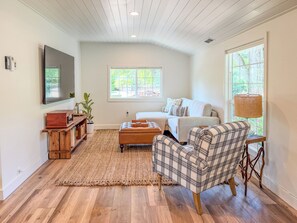 A living room with a sofa, ottoman, and chair, a flat-screen TV, and a large window with a view of greenery. - The Little House on Rocky Hill - Peaceful 2BR Newberg Wine Country Retreat (Newberg)