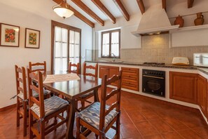 A kitchen with wooden cabinets, a stove, and a dining area with chairs and a table. - Tranquil Pollensa Villa for 5 (Pollensa / Pollença)