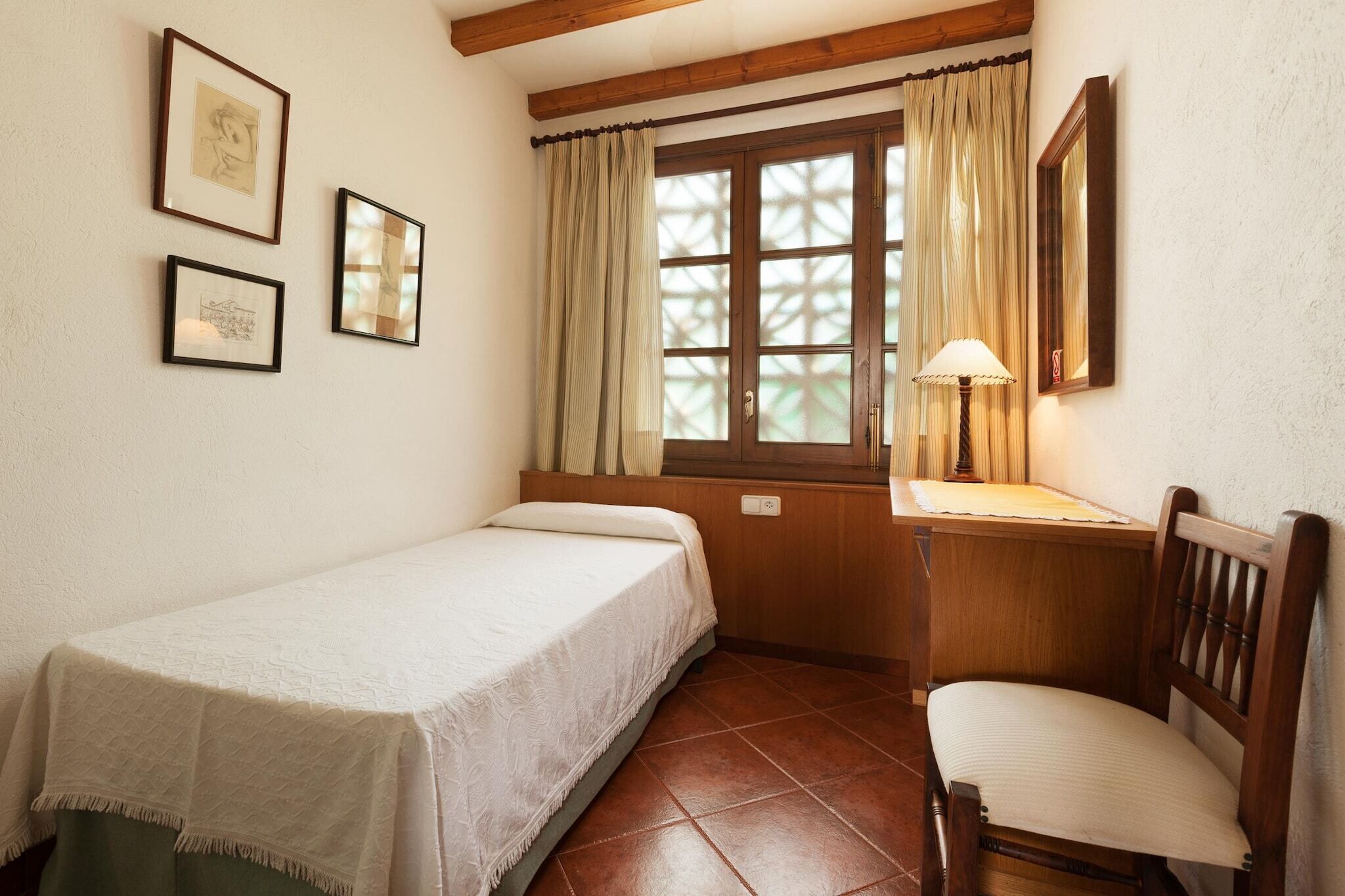 Une petite pièce agréable et chaleureuse, avec un lit simple, un bureau avec une chaise, une fenêtre avec des rideaux et des tableaux encadrés accrochés au mur.