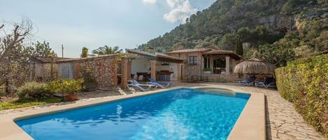 A swimming pool with lounge chairs, a stone building, and a mountain backdrop.