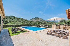 Ein Schwimmbecken mit Liegestühlen und einem Sonnenschirm, umgeben von einem angelegten Garten und Blick auf einen Berg.