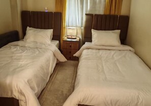 Een hotelkamer met twee enkele bedden, een nachtkastje met een lamp en een raam met gordijnen.