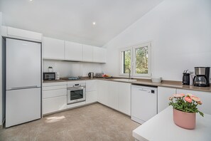 A modern kitchen with white cabinets, a built-in oven, a dishwasher, and a window with a view of greenery. - Premium Pollensa Country Villa for 6 (Pollensa)