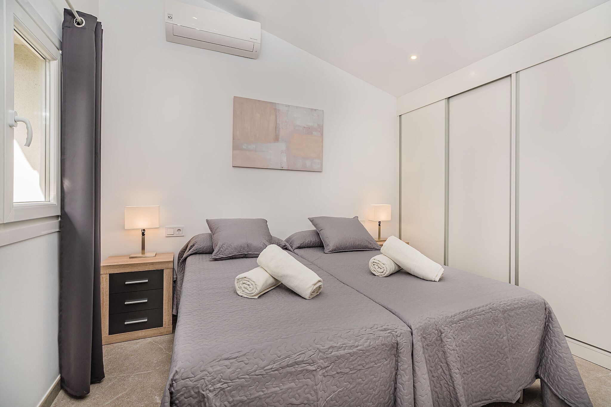 A modern bedroom with two beds, bedside lamps, a wall-mounted air conditioner, and a large abstract painting.