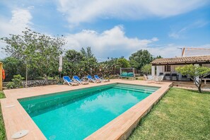 A rectangular swimming pool with clear blue water, surrounded by a wooden deck. There are several lounge chairs and a small table with chairs. The pool area is adjacent to a grassy lawn and a building with a tiled roof. - Romantic Pollensa Country Escape (Pollensa / Pollença)