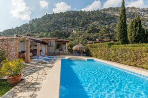 Ein Poolbereich mit Liegestühlen, ein Steinhaus und eine gebirgige Kulisse.