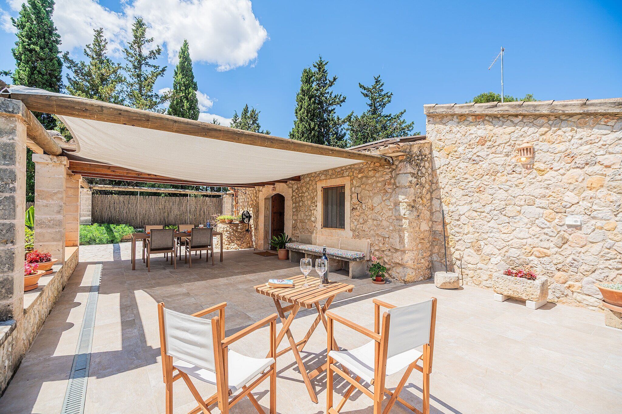 An outdoor patio area with a stone wall, a pergola providing shade, and a seating arrangement with tables and chairs.