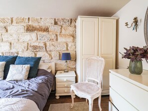 Ein Schlafzimmer mit Stehwand, einem Bett mit blau-weißer Bettwäsche, einer weißen Kommode, einem Korbstuhl und einem Nachttisch mit Lampe.
