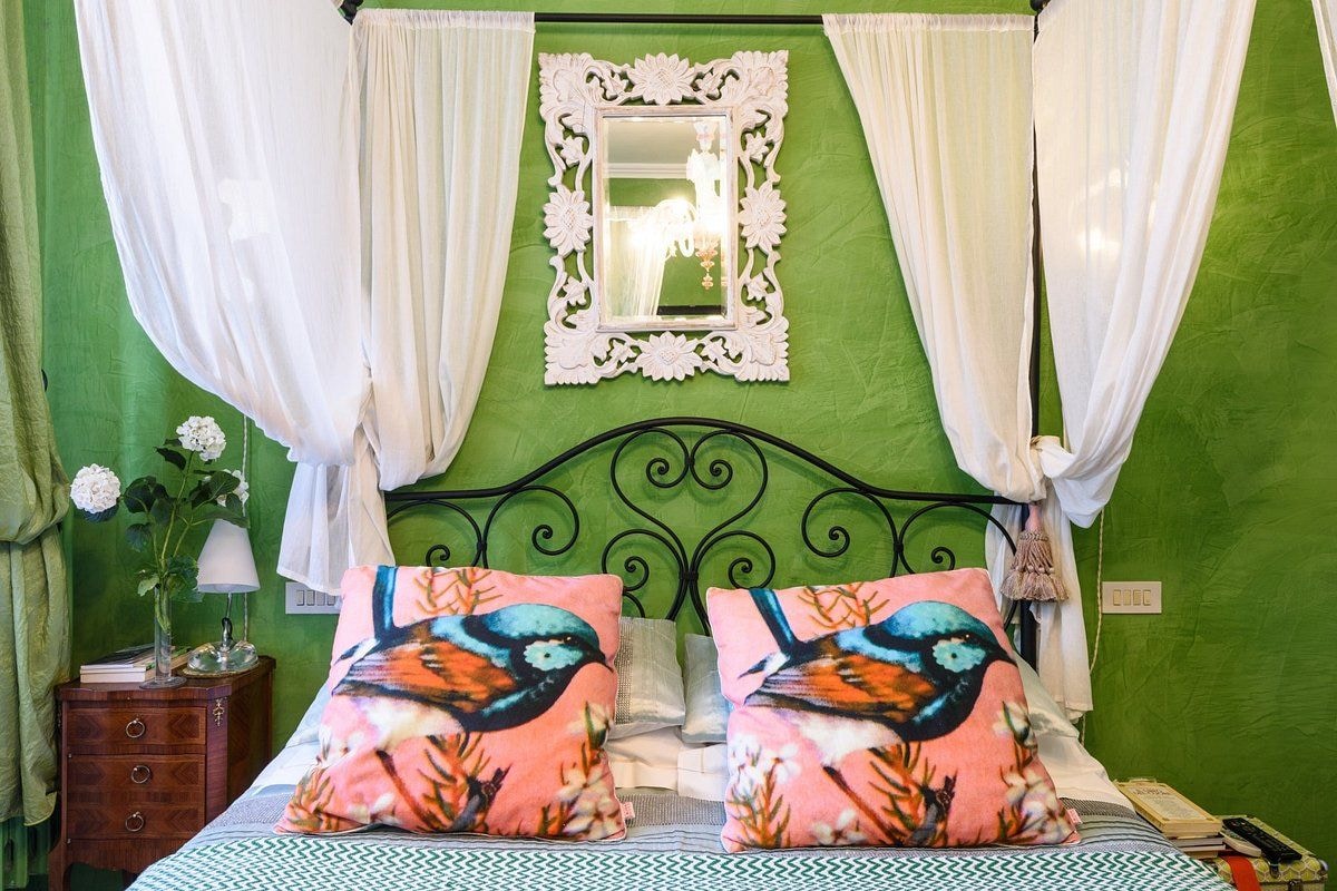 Quarto com parede verde, cama com duas almofadas com desenho de pássaro, espelho com moldura floral e mesa de cabeceira com luminária e flores.