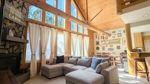 A cabin interior with a large L-shaped sectional sofa, a stone fireplace, and a kitchen area. - Sage Creek Chalet At The Running Y (Klamath Falls)
