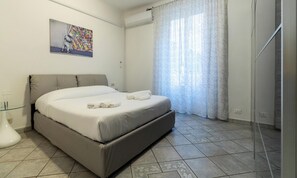 Ein Schlafzimmer mit einem Bett, einem Glastische, einem Bild an der Wand und einem Fenster mit Vorhängen.