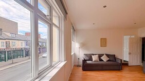 A spacious living room with a large window, a sofa, and a lamp. - Prince Of Wales Rd - Apartment 1 (Norwich)