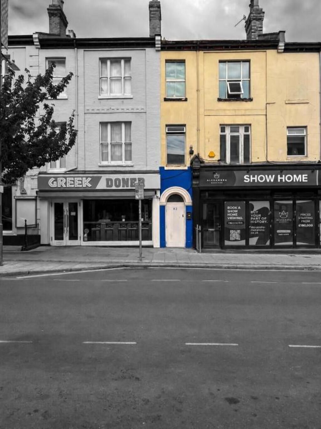 A street view with three commercial buildings: a Greek diner, a blue storefront, and a yellow building with a sign reading 'SHOW HOME'.