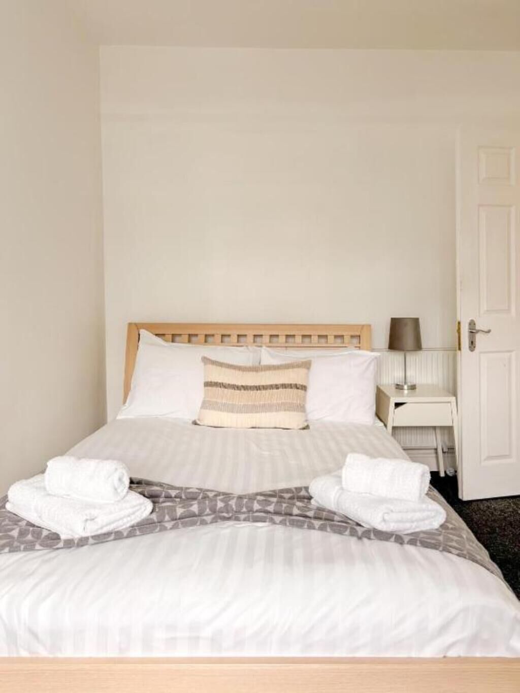 A neatly made bed with a wooden headboard, a bedside table with a lamp, and a door to the right.