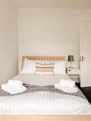 A neatly made bed with a wooden headboard, a bedside table with a lamp, and a door to the right.