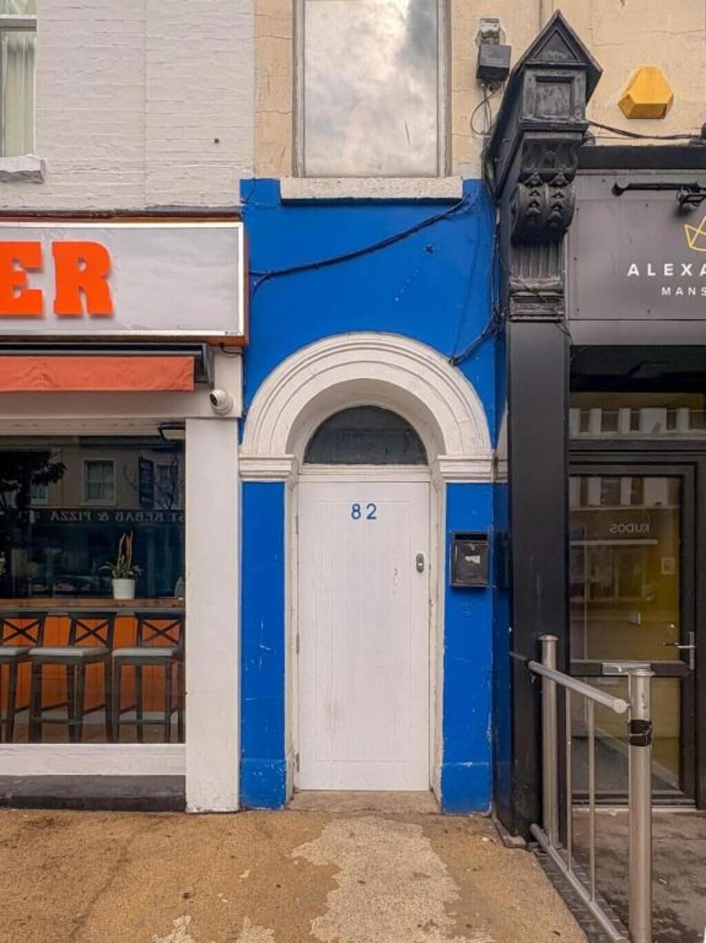 A building facade with a blue and white color scheme, a white door with the number 82, and a visible storefront with a sign that is partially obscured.