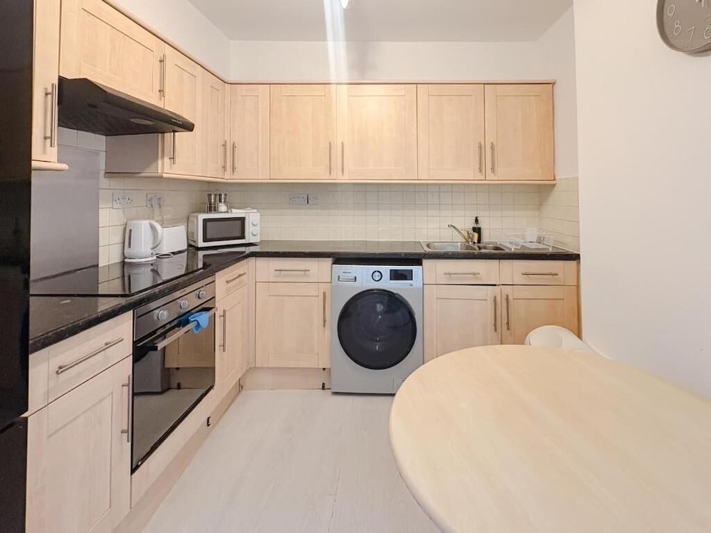 A compact kitchen with wooden cabinets, a microwave, a washing machine, and a dining area with a table.