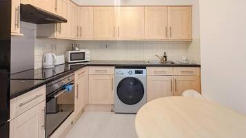 A compact kitchen with wooden cabinets, a microwave, a washing machine, and a dining area with a table.