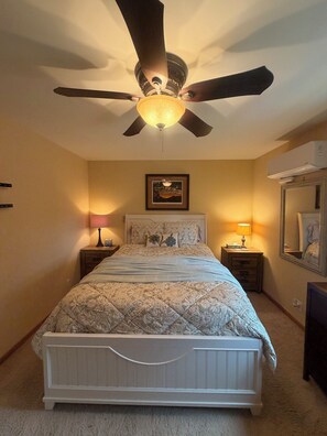 A bedroom with a bed, two bedside tables, a ceiling fan, a wall-mounted air conditioner, and a framed picture above the bed. - The Rock House of Herron Island (Home)