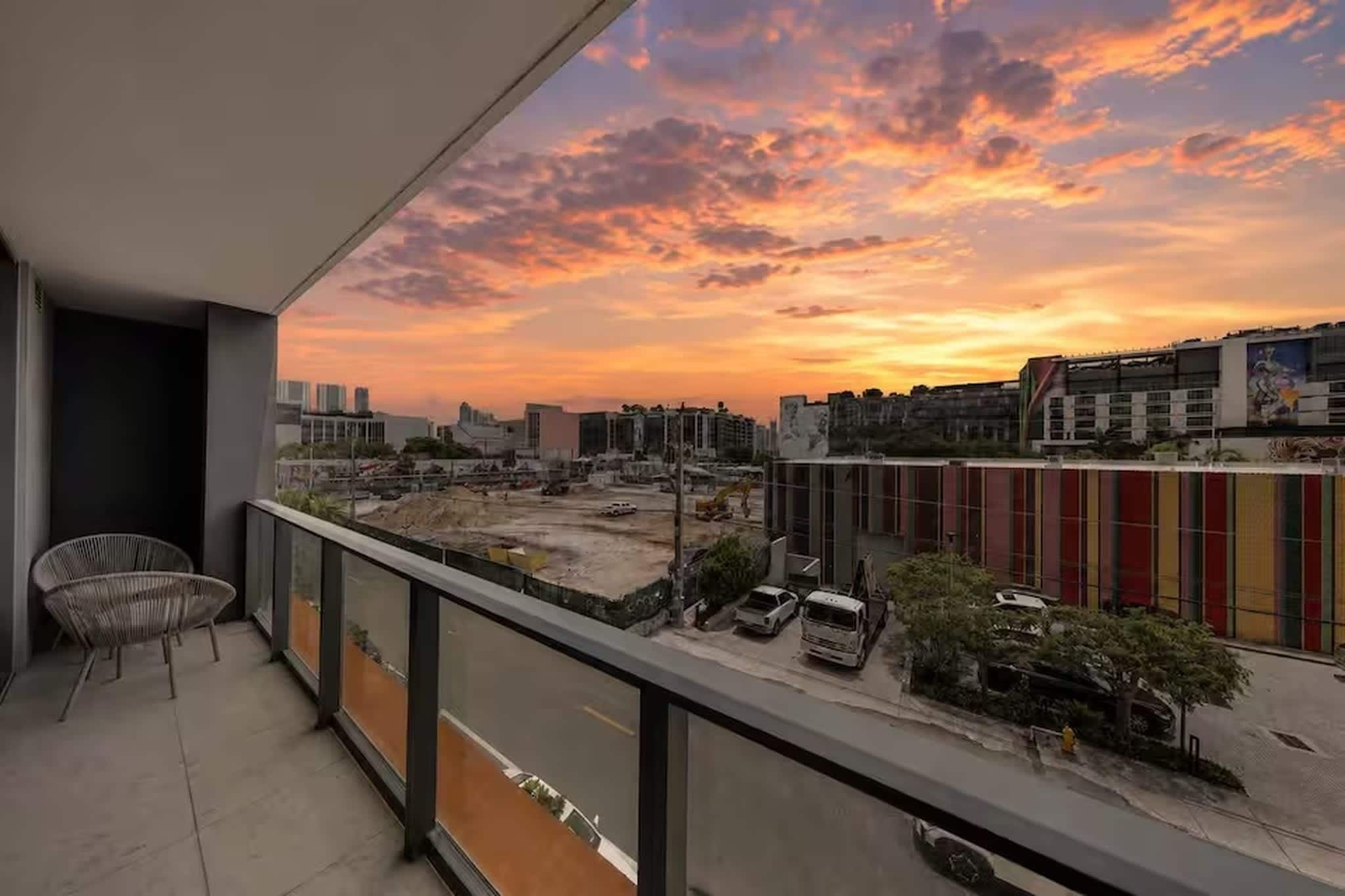 Un balcón con vista a la puesta de sol sobre un paisaje urbano, con edificios y obras de construcción.