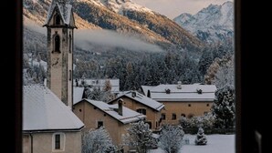 A church with a tall steeple, surrounded by snow-covered buildings and mountains in the background. - Haute Alpine Living (S-chanf)