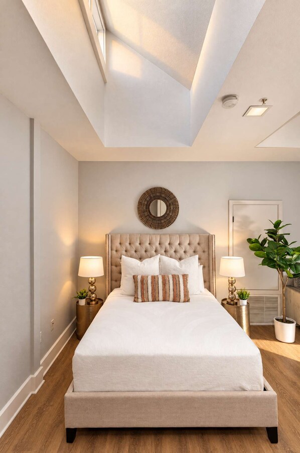 A bedroom with a large bed, two bedside lamps, a round mirror, a potted plant, and a ceiling light. - The Penn Hotel - Smart TV, AC & Downtown Location! (San Diego)