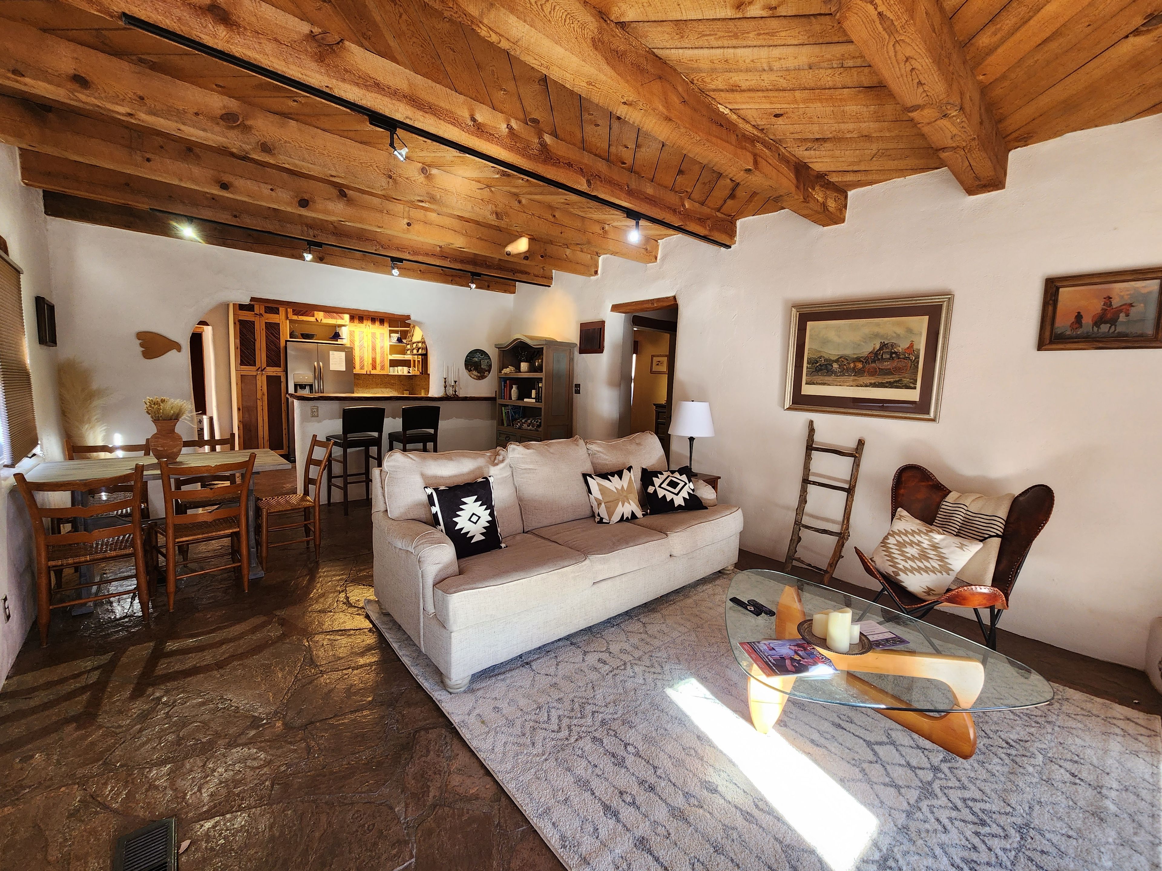 A cozy living room with a wooden ceiling, a sofa with patterned cushions, a glass coffee table, and a dining area with wooden chairs.