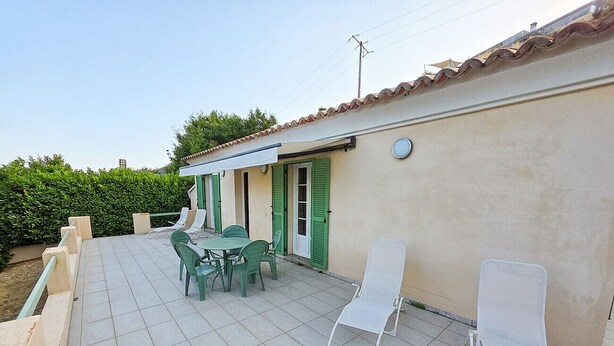 A patio with a table and chairs, a white lounge chair, and a building with green shutters.