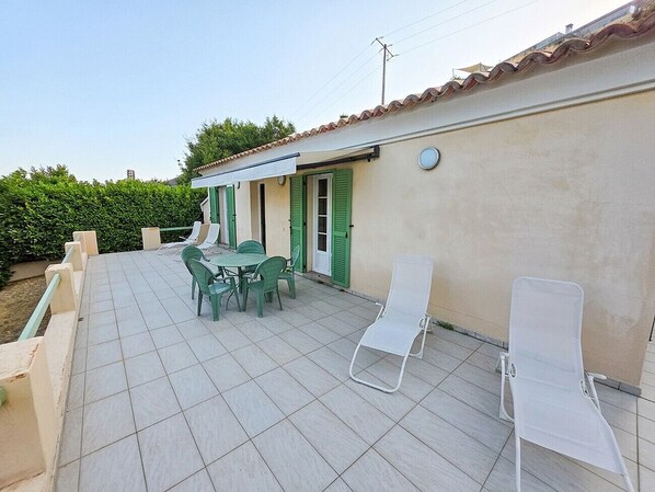 Une terrasse avec une table et des chaises, un fauteuil de jardin blanc et un bâtiment aux volets verts.