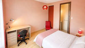 A bedroom with a bed, a desk with a chair, a red chair, a red lamp, and a mirror.