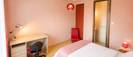 A bedroom with a bed, a desk with a chair, a red chair, a red lamp, and a mirror.