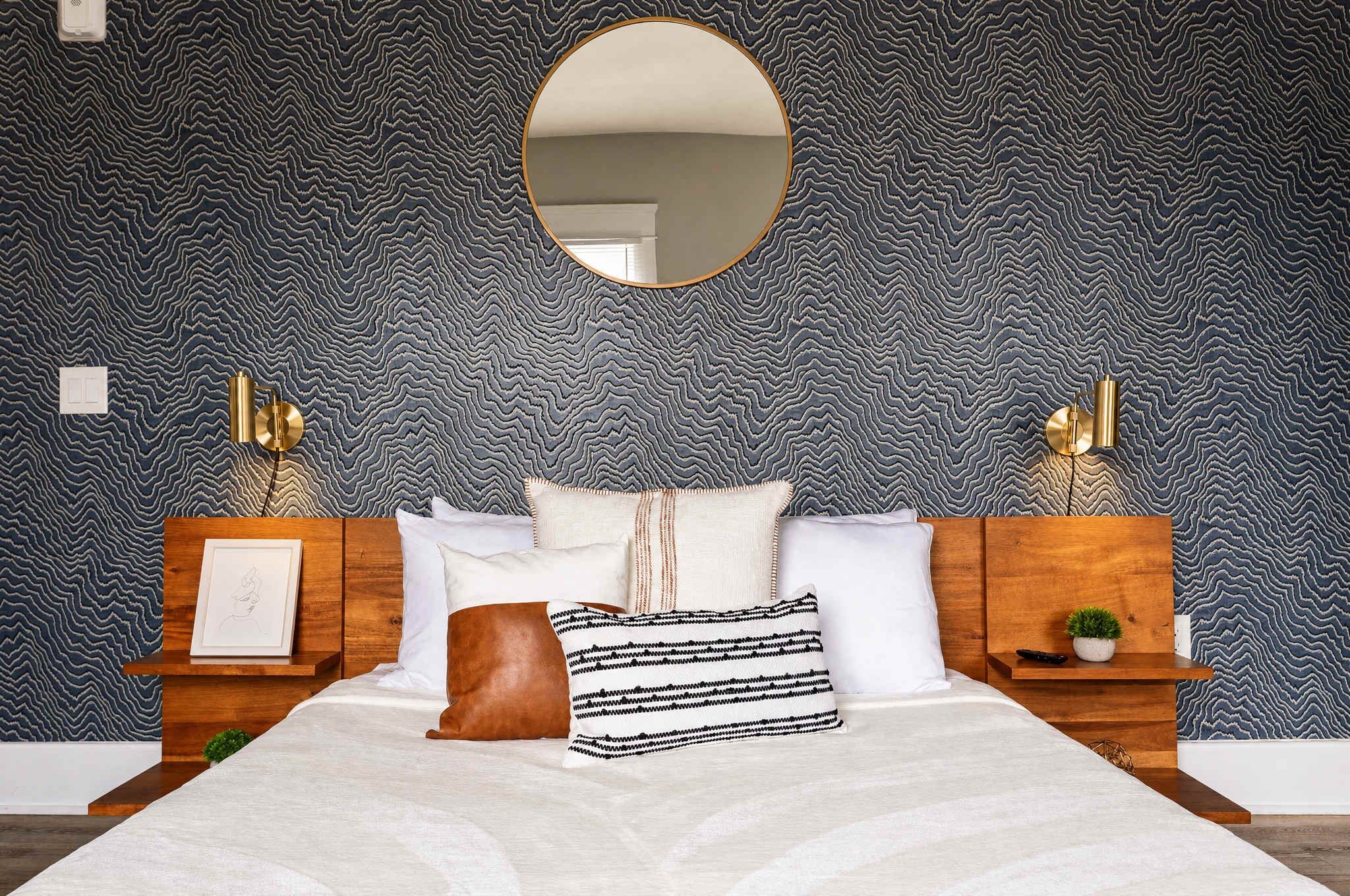 A bedroom with a patterned wallpaper, a round mirror, a bed with multiple pillows, bedside tables with lamps, and a framed picture on the left side.