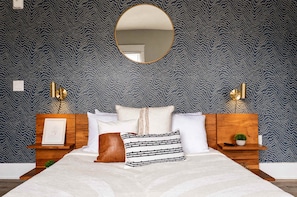 A bedroom with a patterned wallpaper, a round mirror, a bed with multiple pillows, bedside tables with lamps, and a framed picture on the left side. - The Penn Hotel - Queen Bed & Downtown Adventures!! (San Diego)