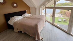 A bedroom with a bed, a large window, and a view of a garden. - Holiday Home 'Beachvilla with Sauna' with Lake View, Wi-Fi and Air Conditioning (Westerhaar-Vriezenveensewijk)