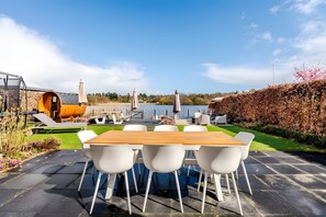 A patio with a wooden table and white chairs, overlooking a lake with a view of a house and trees in the background. - Holiday Home 'Beachvilla with Sauna' with Lake View, Wi-Fi and Air Conditioning (Westerhaar-Vriezenveensewijk)