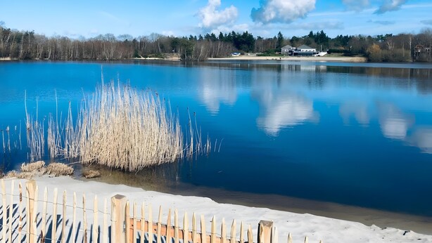 A tranquil lake with a wooden fence in the foreground, a clear sky, and a distant building. - Holiday Home 'Beachvilla with Sauna' with Lake View, Wi-Fi and Air Conditioning (Westerhaar-Vriezenveensewijk)