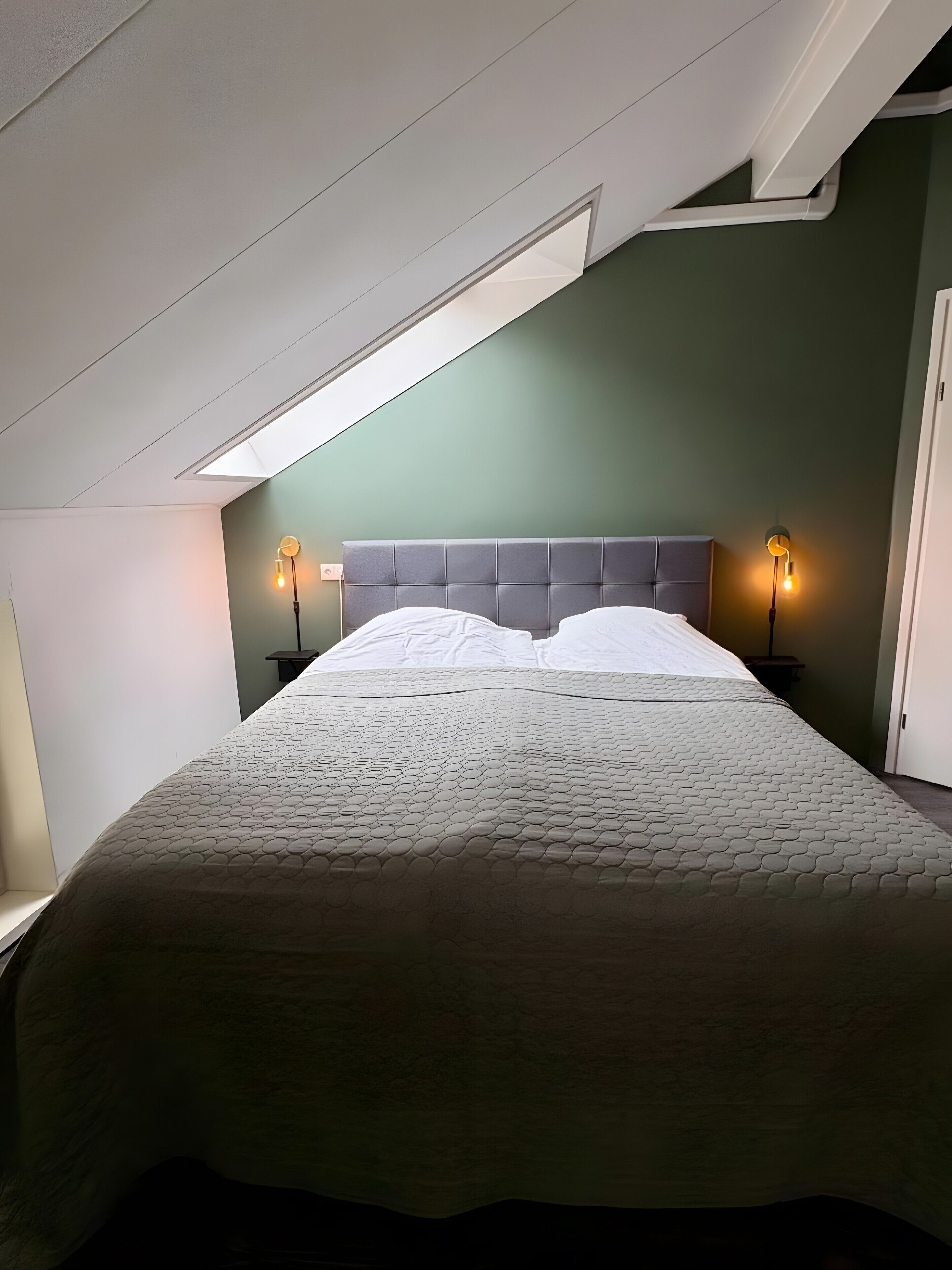 Een slaapkamer met een bed, twee bedlampen, een schuine balkenkap en een raam.