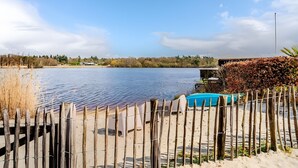 A lakeside view with a wooden fence, a blue pool, and a building in the background. - Holiday Home 'Beachvilla with Sauna' with Lake View, Wi-Fi and Air Conditioning (Westerhaar-Vriezenveensewijk)