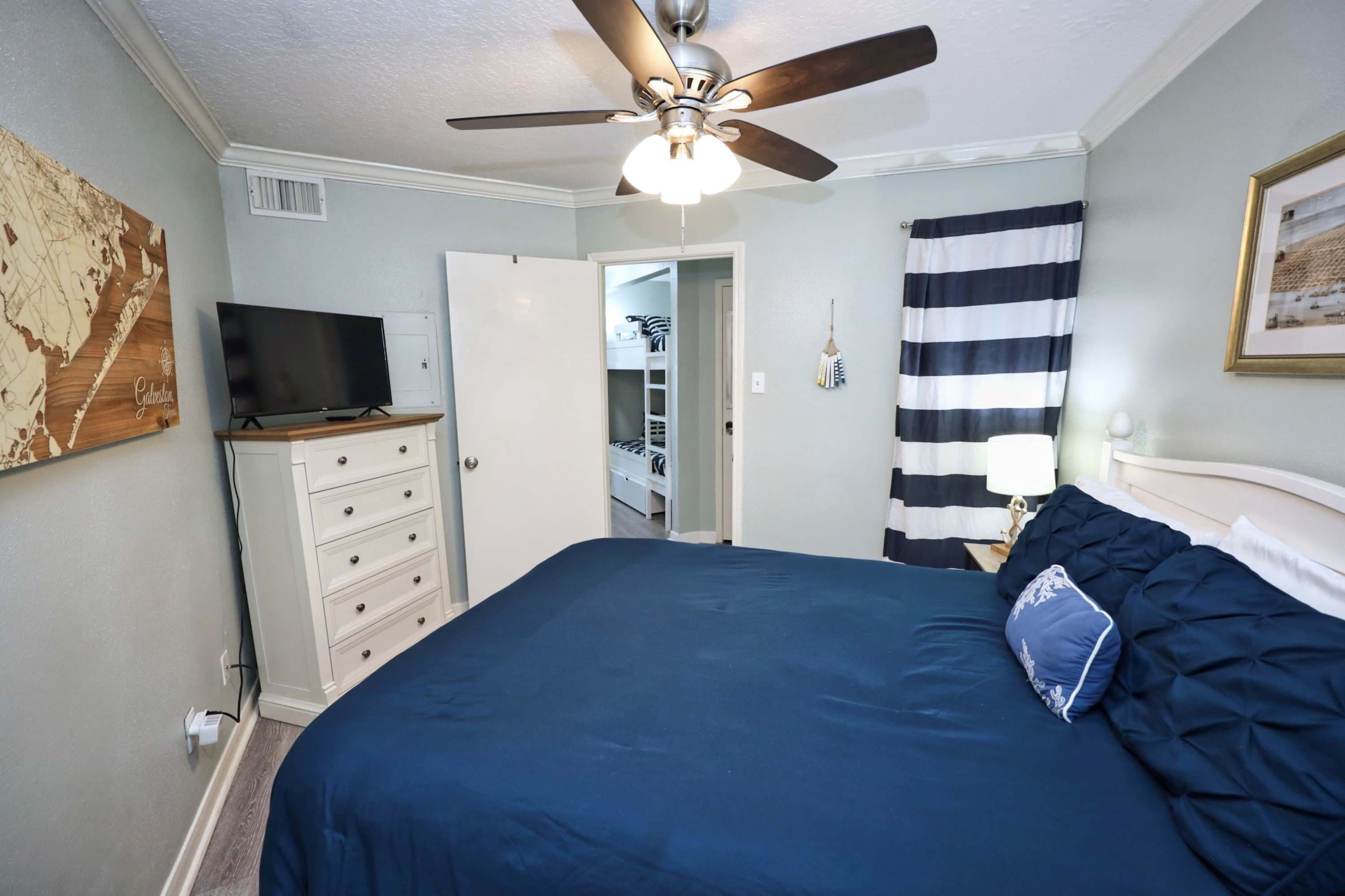 A bedroom with a bed, a ceiling fan, a dresser, a television, and a striped curtain.