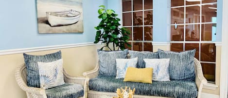 Una habitación con pared azul claro, un cuadro de un barco, una planta grande en maceta, muebles de mimbre y una mesa de centro de vidrio con una bandeja de aperitivos.