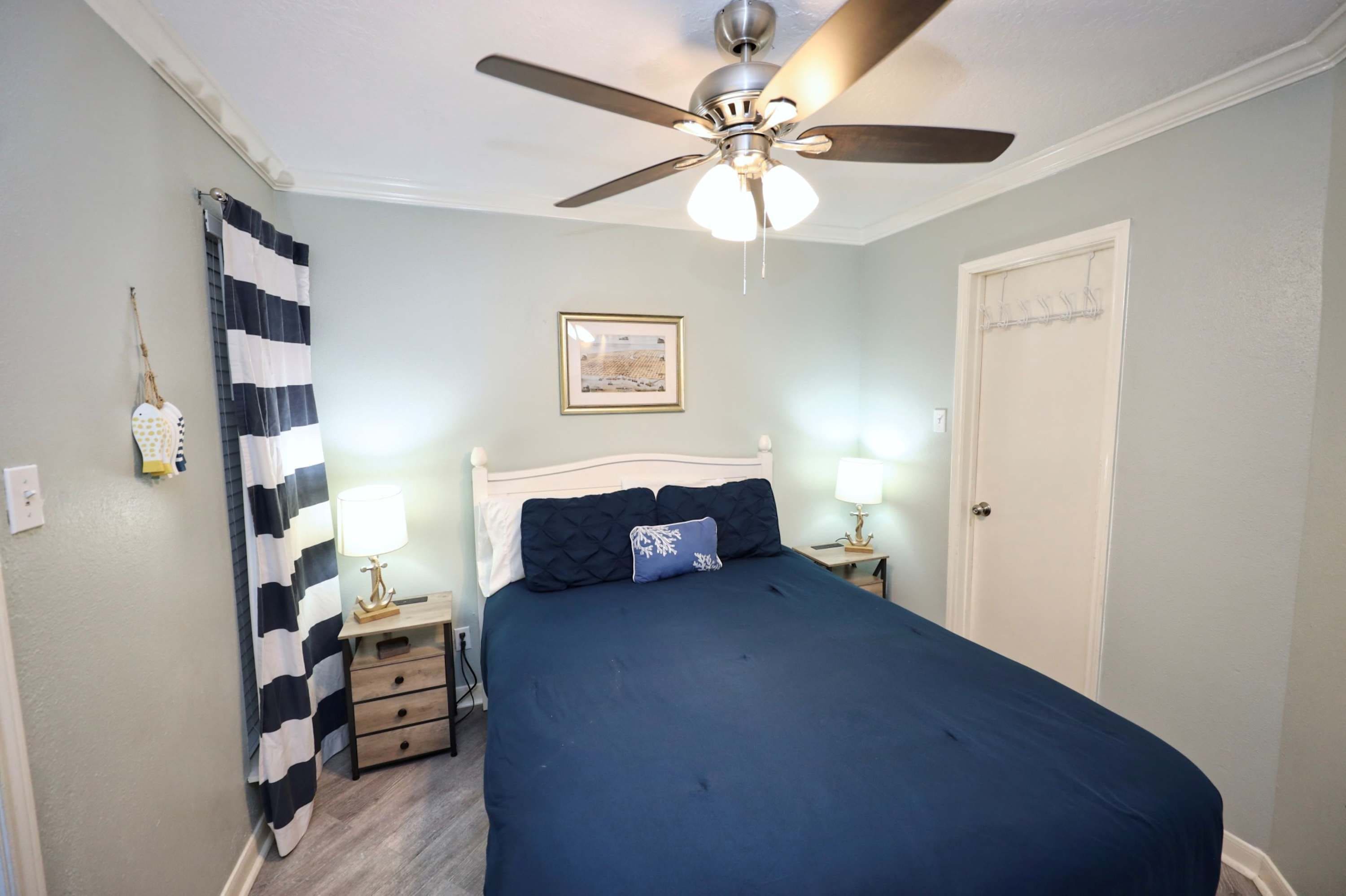 Un dormitorio con cama, ventilador de techo, mesita de noche con lámpara, una puerta y una lámpara fijada a la pared.