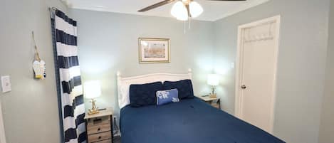 Un dormitorio con cama, ventilador de techo, mesita de noche con lámpara, una puerta y una lámpara fijada a la pared.