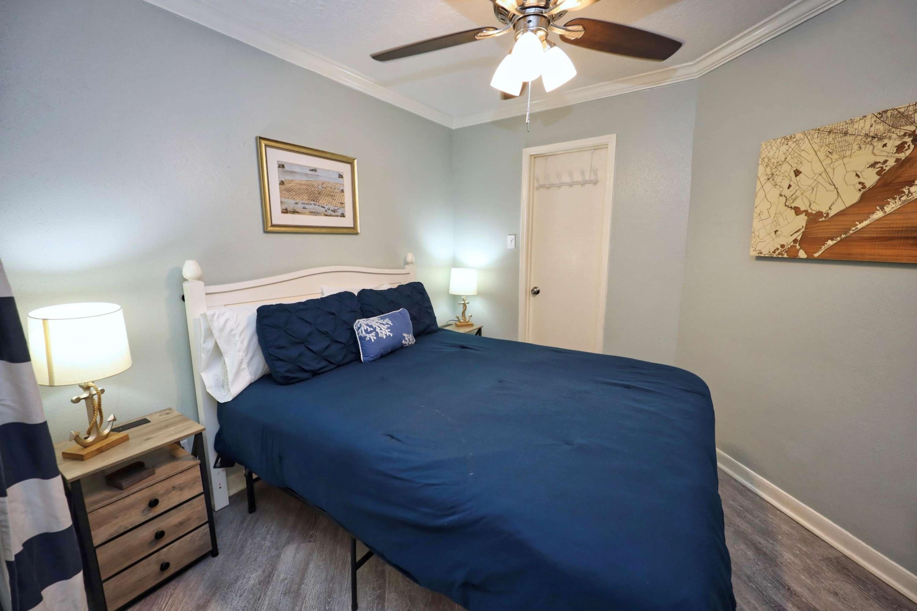 A bedroom with a bed, a ceiling fan, a nightstand with a lamp, a dresser, and a door.